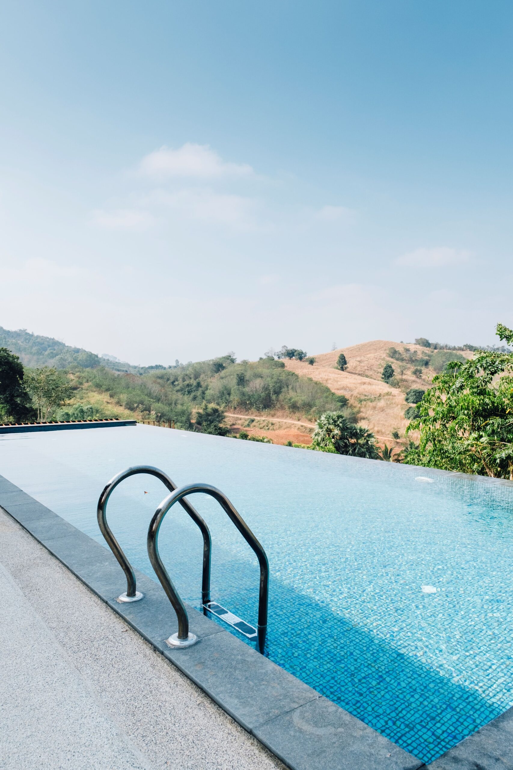 Piscina a sfioro con scaletta in acciaio, affacciata su colline verdi e dorate sotto un cielo azzurro con leggere nuvole. 