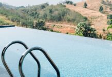 Piscine a sfioro: il fascino dell’acqua che si fonde con l’orizzonte Piscina a sfioro con scaletta in acciaio, affacciata su colline verdi e dorate sotto un cielo azzurro con leggere nuvole.