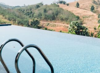 Piscine a sfioro: il fascino dell’acqua che si fonde con l’orizzonte Piscina a sfioro con scaletta in acciaio, affacciata su colline verdi e dorate sotto un cielo azzurro con leggere nuvole.